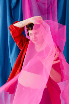 Young curvy non binary person standing behind pink veil