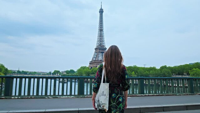 Beautiful girl in a floral dress walks to the Tower with local bag with Paris logo. Young woman explores the Capital of France with the historical Tour in Paris. 