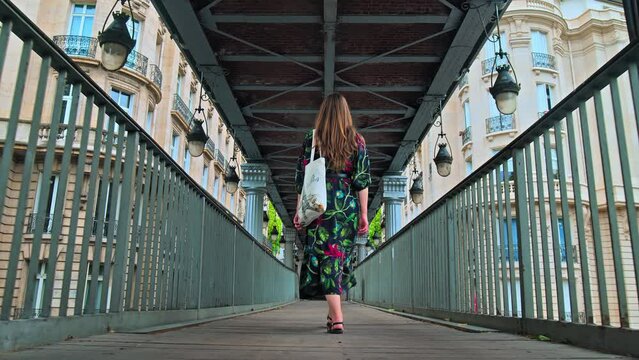 Beautiful girl in a floral dress walks on a bridge with local bag with Paris logo. Young woman explores the Capital of France with old historical buildings and street lamps in Paris. 