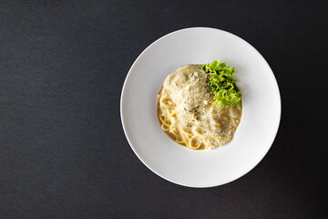 Top view of spaghetti carbonara in a round white plate with green lettuce. Placed on the table, the floor is a black blur polished steel pattern. There is a copy space area on the left side of image.