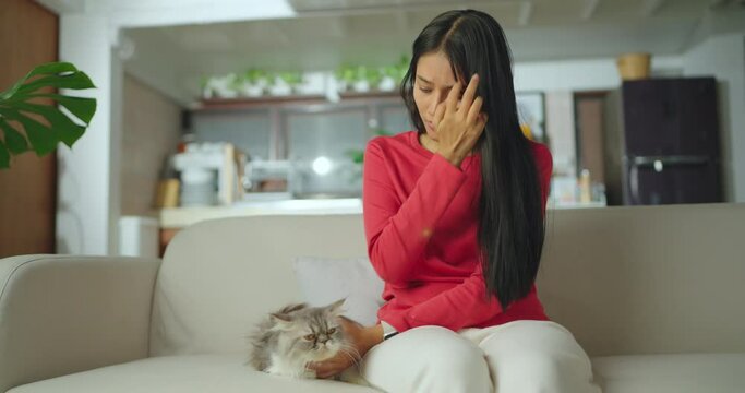 Young asian woman sitting with her cat at living room. Girl hand in sneezing from fur allergy while playing with her lovely cat, pet on carpet in living room at home.