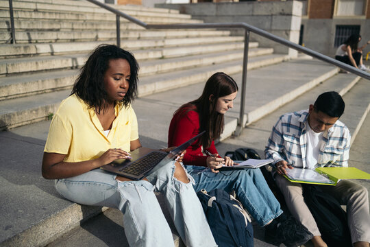 College Friends Sitting On Stairs And Working On Laptop