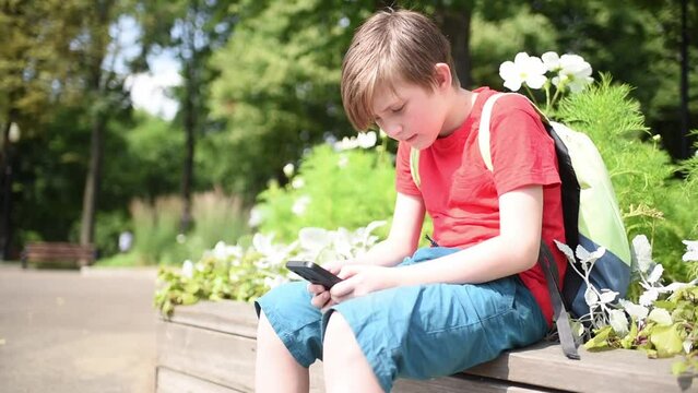 a boy, a schoolboy holds a smartphone in his hands, plays games, surfs the internet in a mobile, sits on a bench in the park near a flower bed on a sunny day