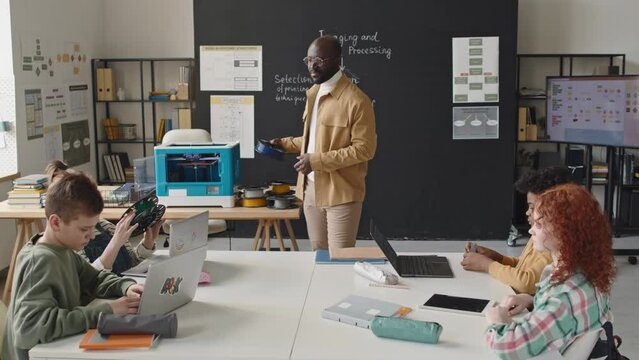 Group of diverse pupils examining filament spool and listening to black male teacher during 3D printing lesson classroom