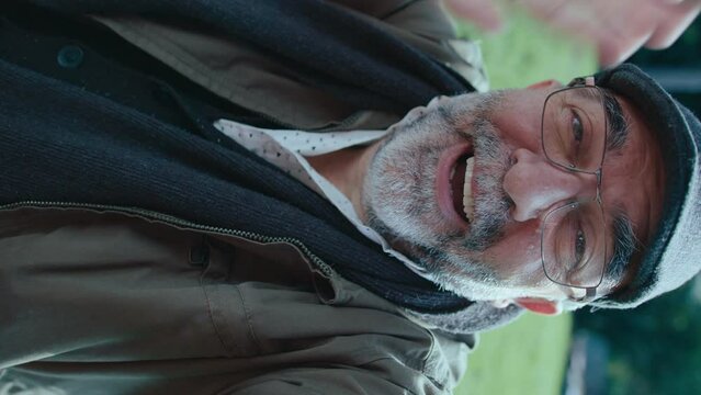 Senior man with gray beard waving and chatting at camera, video calling on a walk outdoors in the park. Vertical format footage, POV shot