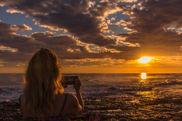 Woman taking photos of the sunset on the beach with mobile phone. Summer, travel and technology...