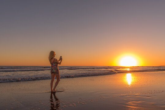 Woman with mobile phone taking photos of sunset standing on the seashore at the beach. Summer, travel and concept.