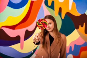 Lovely woman posing covering eyes round lollipop at colored wall, pensive looking at camera. Pretty lady in beige suit showing candy on stick. Summer fashion sweet concept. Copy ad text space poster