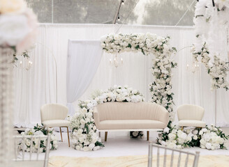 Beautiful decor for the wedding ceremony in the form of a sofa and white flowers on the wedding arch. Light furniture in the form of a sofa and chairs in a white tent is decorated with white roses
