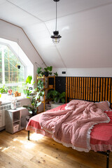 Unmade bed with red striped linen and a mess in Loft style bedroom interior, black wall with wooden slats, metal bed, potted plants on a trapezoidal window in the attic. Modern Green House