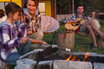 A girl in a plaid shirt roasts marshmallows on a fire in the yard of the house, father plays the guitar. Evening family get-together by the campfire, outdoor picnic in tourist chairs