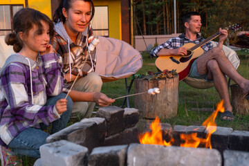 A girl in a plaid shirt roasts marshmallows on a fire in the yard of the house, father plays the guitar. Evening family get-together by the campfire, outdoor picnic in tourist chairs