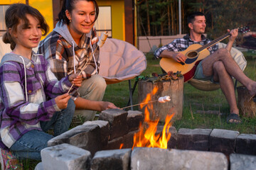 A girl in a plaid shirt roasts marshmallows on a fire in the yard of the house, father plays the...