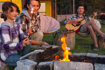 A girl in a plaid shirt roasts marshmallows on a fire in the yard of the house, father plays the guitar. Evening family get-together by the campfire, outdoor picnic in tourist chairs
