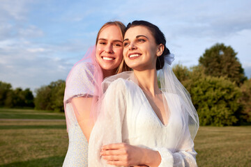 Obraz premium Happy lovely bride in white dress with white veil and bridesmaid in blue dress with pink veil in summer park. Young charming girls smile and hug. Cheerful bachelorette party. Close-up portrait.