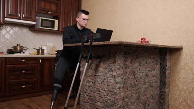 artificial limb, laptop work, assistive solutions. Man with prosthetic leg and glasses works on laptop, crutches close by.