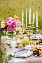 Romantic table decor for a loving couple on a meadow. Two glasses of wine, flowers in a vase, silverware, fruits, chandelier with burning candles, wooden furniture. Sunset, golden hour, summer