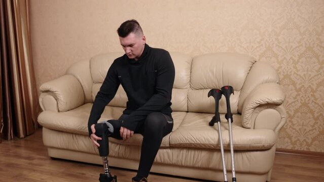 crutches resting, physical impairment, disability technology. Man with prosthetic leg is dressing himself by putting on shoes, seated on couch with his crutches close at hand due to disability.