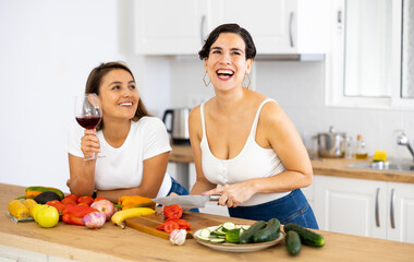 Obraz na płótnie Canvas Happy carefree young latin american lesbian couple chatting carelessly and drinking wine while preparing vegetable salad for dinner in cozy kitchen interior at home
