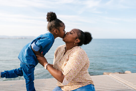 Black mother and daughter kissing near ocean on sunny day