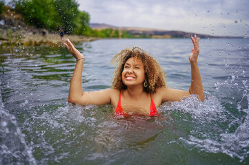 Attractive happy plus size African American Black woman enjoys swimming having fun with water splashes in sea lake water in cloudy weather outdoor