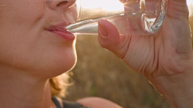 A woman drinks  water from a clear glass on a hot, sunny evening. Slow sips close-up