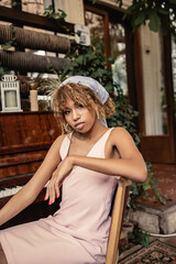 Fototapeta premium Portrait of young and stylish african american woman in dress looking at camera while sitting near piano and plants in blurred indoor garden, woman in comfortable and trendy summer outfit