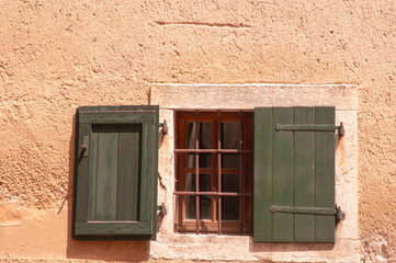 Front view, medium distance of, a  pair of green, wood shutters, one open and one closed, covering barred window