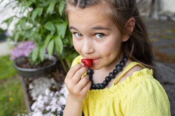 A girl in yellow sits near a flower bed in homemade blueberry beads and eats ripe strawberries