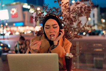 A hijab woman enjoying a sweet dessert at a coffe while working on her laptop during the nighttime in a bustling urban setting.