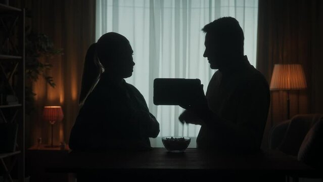 A couple are standing at the table watching a photo, video, film on a tablet. They discuss what they saw, eat popcorn, throwing popcorn. The dark silhouette of a married couple in the living room.