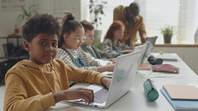 Portrait of African American school boy using laptop at desk in classroom and then smiling on camera during programming lesson, classmates and teacher in background