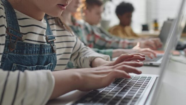 Tilt down shot of little girl typing on laptop at desk while coding during computer class in primary school