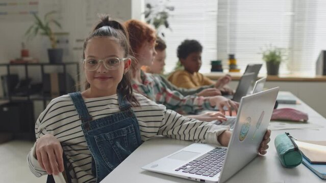 Portrait of cheerful school girl coding on laptop at desk in classroom and then smiling on camera during computer class