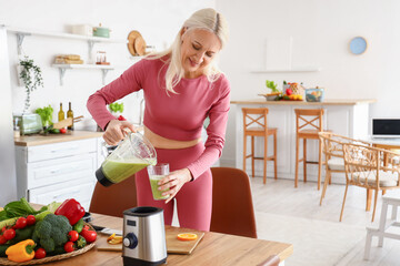Sporty mature woman pouring healthy smoothie into glass in kitchen