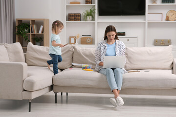 Little daughter playing while her mother working remotely on sofa at home