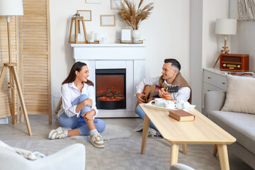 Young woman and her husband playing guitar near fireplace at home