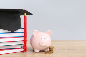Piggy bank, stack of coins, books and graduation hat on wooden table against grey background....