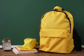 Yellow school backpack with alarm clock, pencil case and notebooks on wooden table near green...