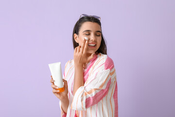 Beautiful young woman applying sunscreen cream on lilac background