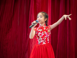 The Asian kid girl sing a song on stage at her school activity day, dress in Qipao style, red curtain background