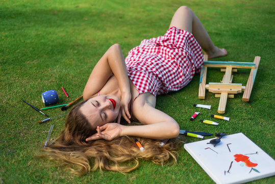 Beautiful woman artist and illustrator laying on the lawn with dropped easel, drawings, pencils