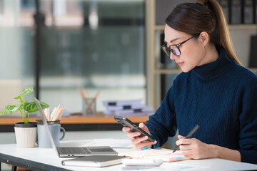 Beautiful Asian woman using a smartphone and working with a laptop while sitting at an office desk, working from home concept.