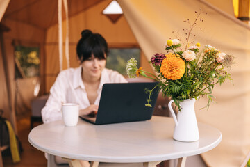 female traveler sitting beside camping tent, drinking morninf coffee and using notebook laptop computer working