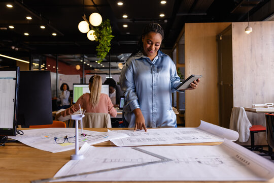 Happy plus size african american female architect using blueprints and tablet in office