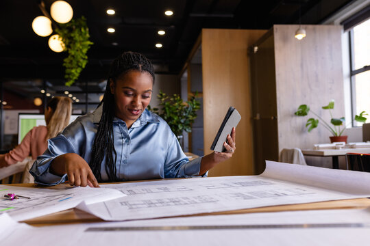 Happy plus size african american female architect using blueprints and tablet in office