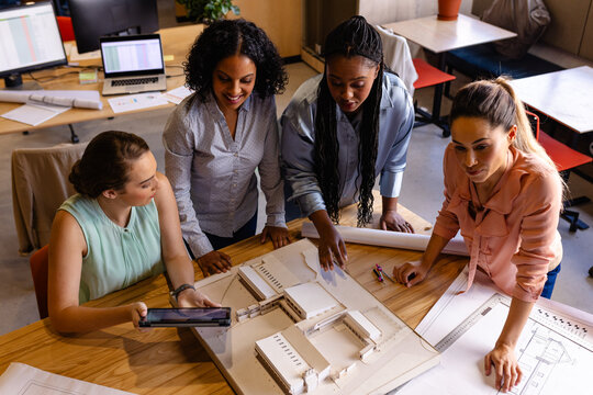 Diverse female architects in discussion using model, blueprints and tablet in casual office meeting
