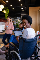 Portrait of happy african american casual businesswomen in wheelchair holding tablet in office