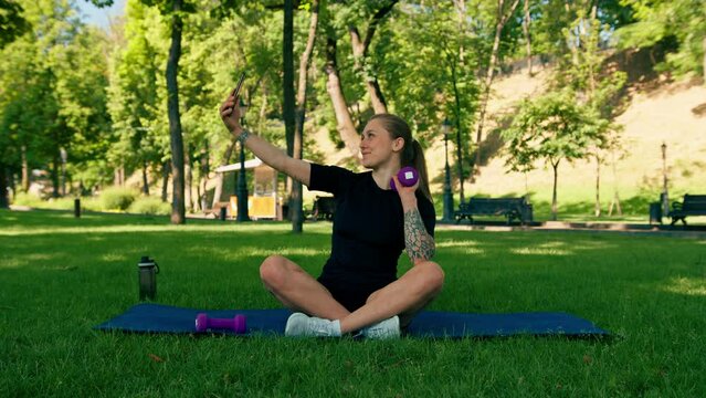Portrait of a young female athlete on a sports mat resting after a workout in the park taking a selfie on a smartphone the concept of a healthy lifestyle