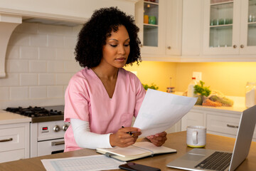 Happy biracial female medical worker in scrubs using laptop working at home in kitchen